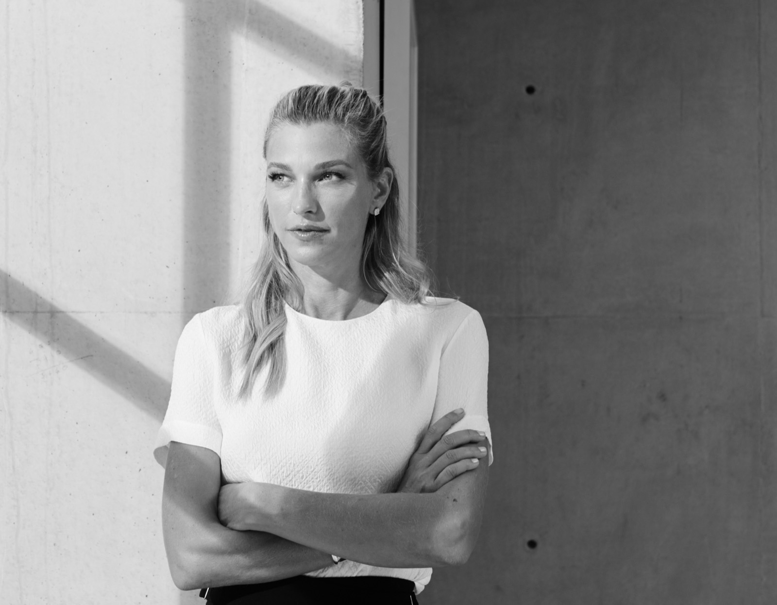 Schwarz-Weiß-Editorialporträt von Nicole Junkermann in weißem Oberteil, mit verschränkten Armen an einer Betonwand im weichen Tageslicht